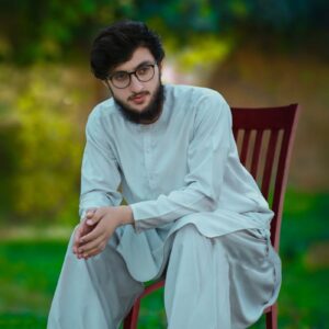 Young man in traditional white shalwar kameez sitting outdoors on a chair.