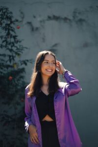 A young woman in a purple shirt smiling and posing outdoors, embracing the natural light.