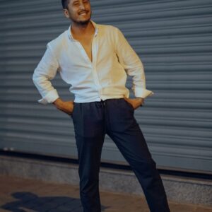 A fashionable man in a white shirt poses confidently on a city sidewalk at night.