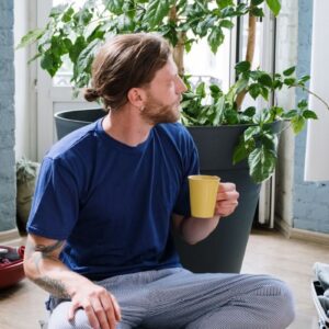 A man enjoying a coffee while sitting in pajamas next to houseplants indoors.